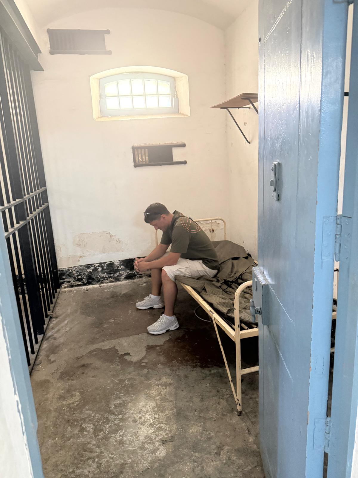 Original location photograph: a young man in everyday clothing sitting on the iron bunk in a real Kordin cell beside the blue doorway