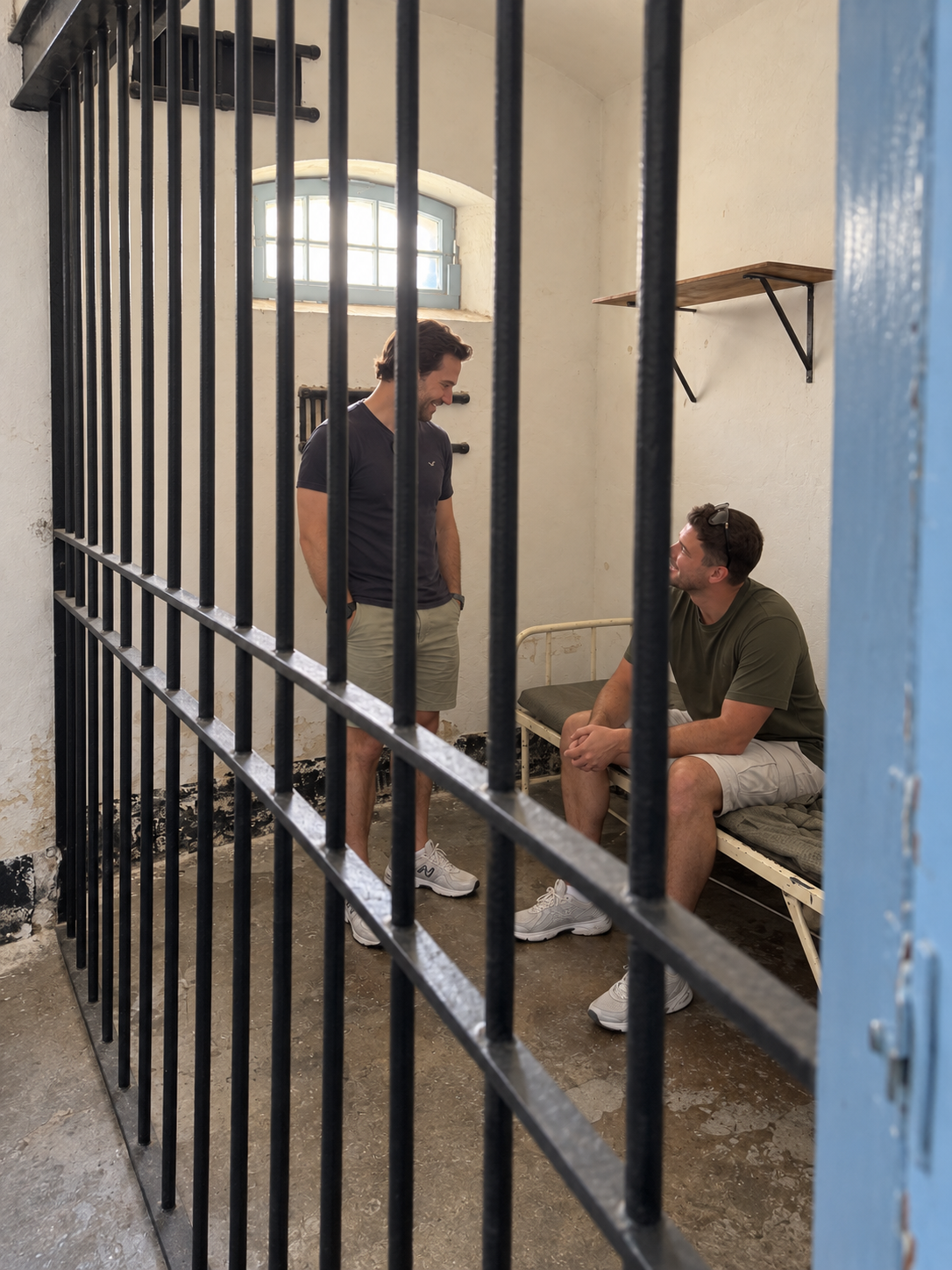 AI-edited concept based on the real Kordin cell: two adult colleagues in everyday clothes, one seated on the bunk and one standing beside him inside the cell
